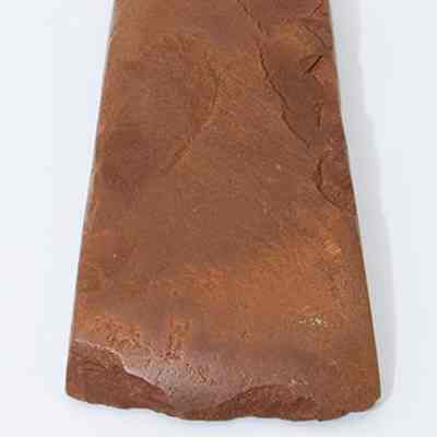 Top lengthwise view of basalt adze with ruler: The image shows a reddish-brown, flat, triangular stone adze on a white surface, with a measuring scale beneath it indicating centimeters and millimeters.