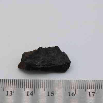 Closeup of basaltic glass flake with ruler: A small, dark basaltic glass flake is placed beside a measuring ruler, highlighting its relatively small size against the neutral background.