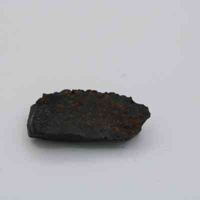 Profile of basaltic glass flake: The image shows a dark, uneven basaltic glass flake resting on a plain white background, highlighting its rough, textured surface.