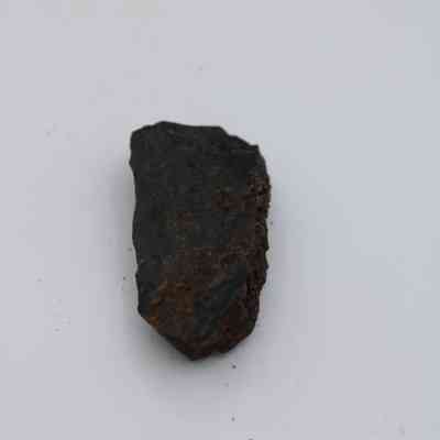 Closeup of basaltic glass flake: The image shows a rough, dark basaltic glass flake resting on a light background, highlighting its rugged texture and irregular edges against the smooth surface.