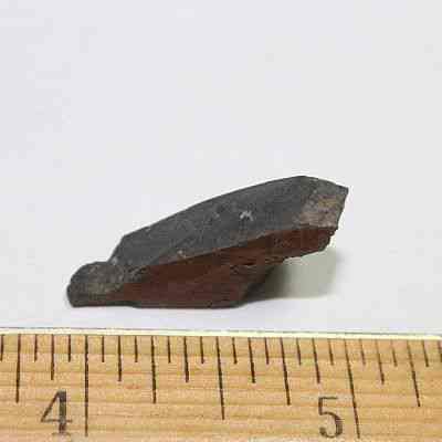 Profile of polished basalt piece: A small, dark, jagged rock fragment is placed against a light background, with a wooden ruler beside it showing its length is approximately one inch.
