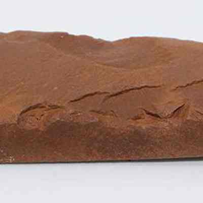 Profile of a polished basalt adze: The image shows a brown, rectangular stone adze with an uneven surface, featuring some small natural ridges and smooth edges, set against a plain white background.