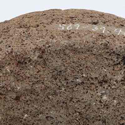 'Ulu maika fragment with visible alternate id number: The image shows a coarse, textured stone with an uneven surface, featuring a light brown hue with darker spots. There is white writing on the top,