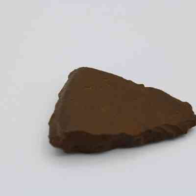 Closeup of small adze fragment: The image depicts a small, triangular fragment of an adze, featuring a dark brown color and a slightly uneven surface, resting on a smooth, white surface.