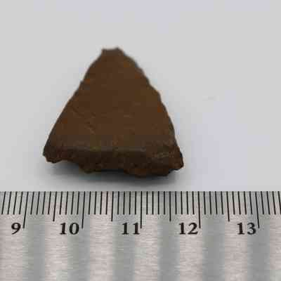 Adze fragment with ruler: The image shows a triangular, brown adze fragment placed beside a metal ruler displaying centimeters, emphasizing its small size and archaeological significance.