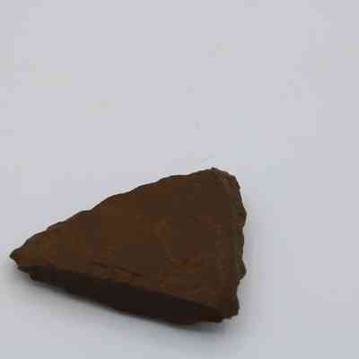 Profile of a small adze fragment: The image shows a triangular fragment of an adze, made of stone with rough, uneven edges, resting on a plain surface.