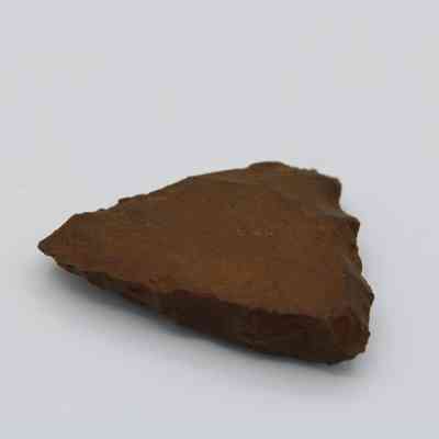 Closeup profile of adze fragment: The image shows a triangular, rough-edged adze fragment resting on a smooth, light-colored surface, showcasing its weathered texture and brown hue.