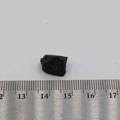 Small basaltic glass flake and ruler: A small piece of basaltic glass flake rests on a surface beside a metal ruler for scale reference, highlighting its texture.
