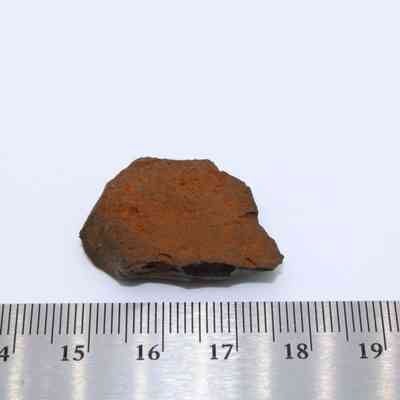 Basaltic glass flake with metal ruler: A small basaltic glass flake, measuring approximately 3 centimeters, displays a rough, rust-colored surface beside a stainless steel ruler.