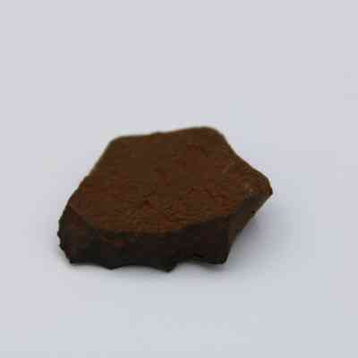 Closeup of basaltic glass flake: The image shows a small, rugged flake of basaltic glass resting on a plain, neutral surface, showcasing its dark, earthy texture.