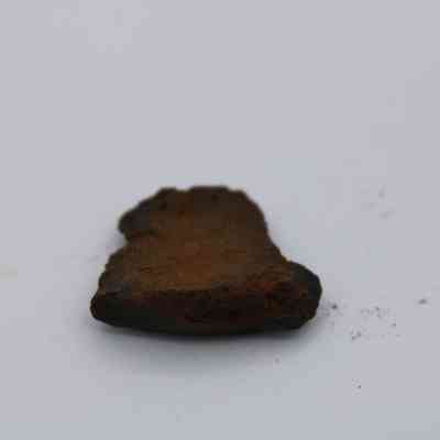 Closeup of basaltic glass piece: The image shows a rough, brownish piece of basaltic glass with uneven edges, resting on a plain white surface, with some scattered dust nearby.
