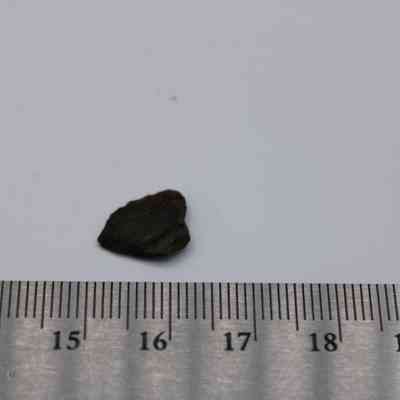 Closeup of basaltic glass flake and ruler: A small, dark flake of basaltic glass rests on a light surface next to a ruler measuring its approximate size.