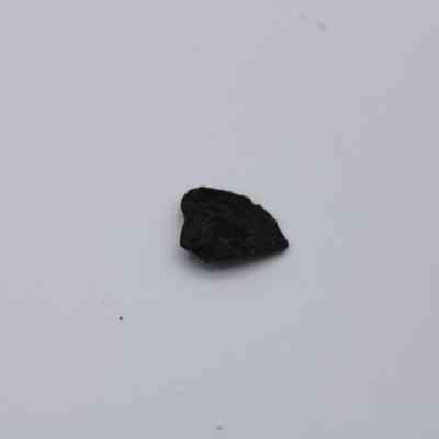 Closeup of tiny basaltic glass flake: A small, dark basaltic glass flake lies on a smooth, light gray surface, showcasing its irregular shape and slightly reflective texture.