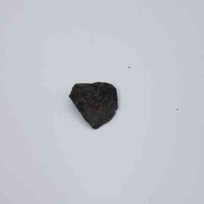 Tiny basaltic glass flake: The image displays a small, dark basaltic glass flake situated against a plain white background, showcasing its rough texture and irregular shape.