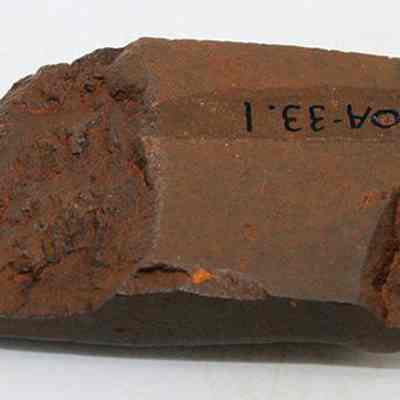 Adze fragment with alternate id number visible: The image shows a weathered, rust-colored fragment of an ancient adze, with visible edges and a code inscribed on its surface.