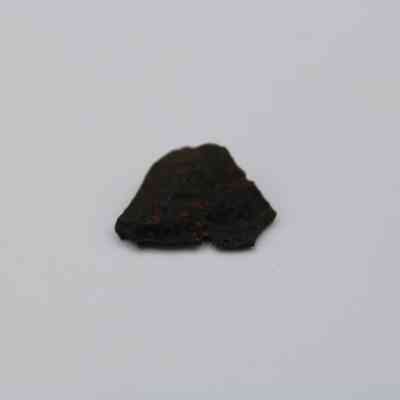 Closeup of basaltic glass flake: A dark, irregularly-shaped basaltic glass flake lies centered against a stark, plain white background, highlighting its textured surface and rugged form.
