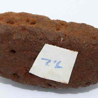 Closeup of base of net sinker with visible alternate id number: The image shows a rough, rust-colored rock sinker with a textured surface, featuring a small piece of white paper attached with a handwritten number.