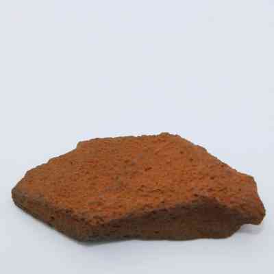 Closeup profile of basaltic glass flake: This image shows a red-brown basaltic glass flake with a rough, porous texture, resting on a plain white background.