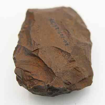 Close up of basalt adze blank with visible alternate id number: The image shows a brown, rough-edged stone tool placed on a light surface.