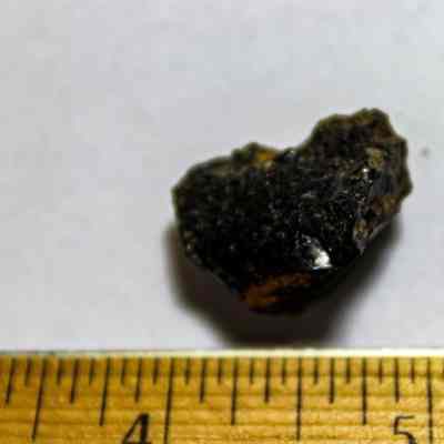 Closeup of a piece of volcanic glass: A small, dark, rough-textured rock is placed on a white surface beside a wooden ruler.