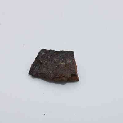 Closeup of basaltic glass flake: A small, dark basaltic glass flake with a rough texture lies against a plain, light-colored background.