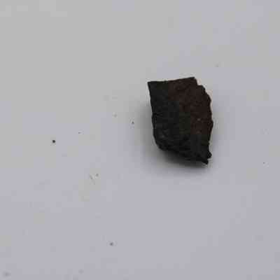 Basaltic glass flake: A dark, irregular basaltic glass flake lies on a smooth white surface, showcasing its rough texture and natural composition.