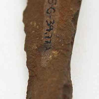 Lengthwise view of basalt with alternate id number visible: The image shows a weathered, ancient stone awl with visible inscriptions or markings carved along its uneven surface, suggesting archaeological significance.