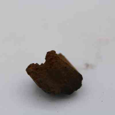 Closeup of basaltic glass core: The image shows a small, brown fragment of basaltic glass core, resting on a plain white surface with a slightly rough texture.