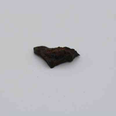 Closeup of basaltic glass flake: The image shows a small, rough, dark brown basaltic glass flake with irregular edges, resting on a plain, light background surface.
