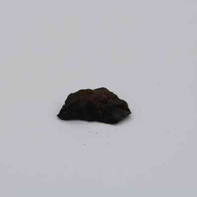Closeup of basaltic glass flake: A small, dark basaltic glass flake is positioned against a plain white background, highlighting its rough, irregular texture and edges.