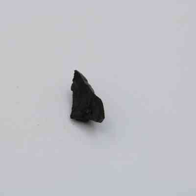 Basaltic glass flake: A small, dark, irregularly-shaped basaltic glass flake rests on a plain, light background, capturing its rugged texture.