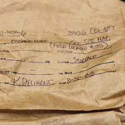 Artifact bag for cowrie lure: The image shows a brown paper bag with handwritten details about a cowrie lure. It includes information on location, surface, and recording date.