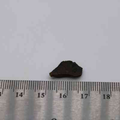 Closeup of basaltic glass flake with ruler: A small, dark basaltic glass flake is placed on a white surface next to a silver ruler for scale measurement.