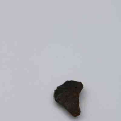 Closeup of basaltic glass flake: A small, dark basaltic glass flake rests on a smooth, white surface, showcasing a rough texture and irregular shape.