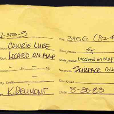 Artifact envelope for cowrie lure: The image shows a yellow envelope with handwritten information detailing a specimen named "Cowrie Lure," located on a map, categorized as surface collection.