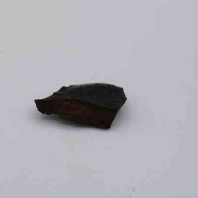 Closeup of basaltic glass flake: The image displays a small, dark basaltic glass flake, showcasing its rough, angular edges against a plain, light background.