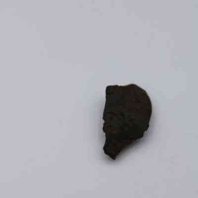 Closeup of basaltic glass flake: The image shows a small, dark basaltic glass flake with a rough texture lying on a smooth, light surface.