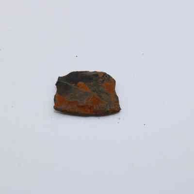Closeup of basaltic glass flake: A small, rust-colored flake of basaltic glass lies on a white surface, displaying a rough texture and irregular shape.