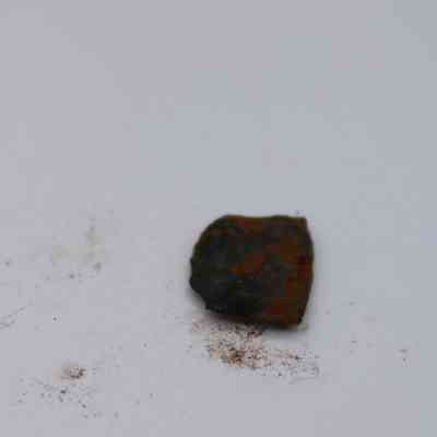 Closeup of basaltic glass flake: The image shows a small flake of basaltic glass resting on a white surface, surrounded by tiny specks of dirt.