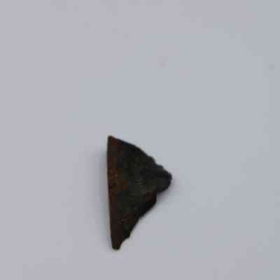 Closeup of tiny basaltic flake glass: The image shows a small, triangular piece of dark basaltic glass flake resting on a plain light background.