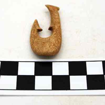 Fishhook: The image shows a small, intricately carved fishhook placed above a black-and-white centimeter scale for measurement reference on a plain background.