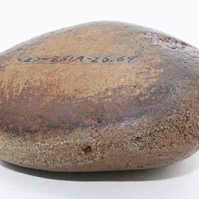 Profile of basalt hammerstone with visible alternate id number: The image shows an oval, smooth brown stone with a slightly rough texture and markings handwritten in black ink on its surface.
