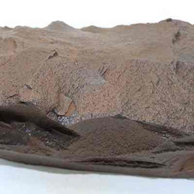 2015.006.2297b: The image shows a large, rough, brown rock adze blank with an uneven texture, displayed against a white background, highlighting its rugged surface details.