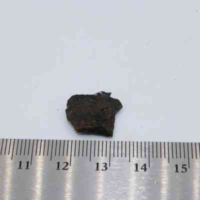 Tight closeup of basaltic glass flake with ruler: A small piece of basaltic glass flake rests on a white surface next to a metal ruler displaying measurements.