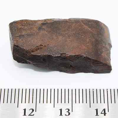 Small adze (ko'i) with a metal ruler: The image shows a dark brown rock adze with rough edges placed above a ruler displaying measurements.