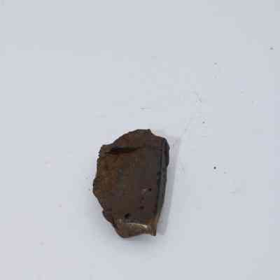 Closeup of basaltic glass flake: The image shows a small piece of basaltic glass flake, characterized by a rough texture and dark coloration, resting on a plain, white background surface