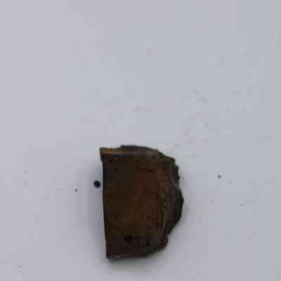 Closeup of basaltic glass flake: A basaltic glass flake lies on a plain surface, displaying a dark, rough texture with an irregular shape and some surrounding specks.