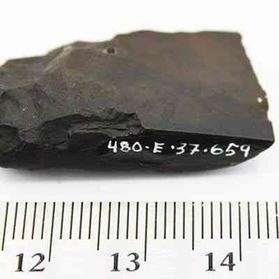 Close up of basalt adze (ko'i) with visible alternate id number and ruler: The image shows a dark, rough-edged rock sample labeled "480-E 37.659" lying above a ruler, measuring approximately 3 cm.