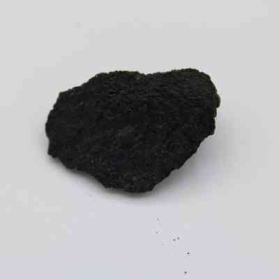 Basaltic glass flake: The image displays a dark, rough-textured basaltic glass flake resting against a plain white background surface.