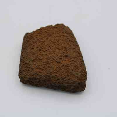 Closeup of basaltic file fragment: The image shows a small, rough-textured fragment of basalt, featuring a dark, porous surface typical of volcanic rock, set against a plain background.